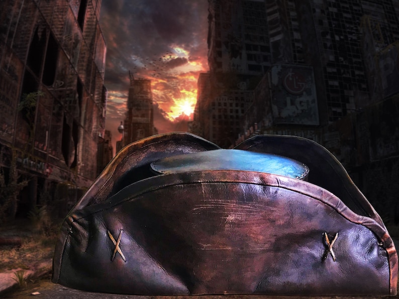 May include: A brown leather pirate hat with a blue lining and two crossed stitches on the side. The hat is lying on a surface in front of a dark, apocalyptic cityscape.