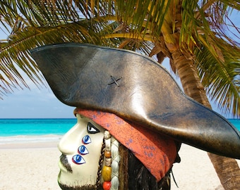 May include: A brown leather pirate hat with a black X stitched on the top. The hat is worn by a person with a white face, red bandana, and three blue eyes painted on the face.