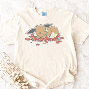 May include: Cream-colored t-shirt featuring a cartoon Eevee character sleeping on red and orange leaves. The Eevee has brown fur, blue ears, and a cream-colored ruff. The shirt has a classic crew neck and short sleeves.