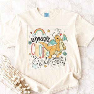 May include: Cream-colored t-shirt featuring a cartoon dragon with teal wings and the phrase "always choose kindness" in colorful letters. The design includes a rainbow, stars, and hearts.