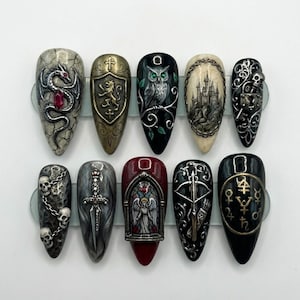 May include: A collection of ten long, pointed false nails featuring gothic and fantasy artwork. Designs include a dragon, a shield, an owl, a castle, skulls, a sword, an angel, a crossbow, and astrological symbols. Each nail has a unique colour and design.