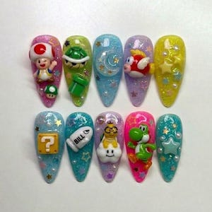 Puede incluir: Un conjunto de diez u&ntilde;as acr&iacute;licas largas en forma de almendra con varios dise&ntilde;os de personajes 3D de Super Mario. Cada u&ntilde;a presenta un personaje diferente, incluyendo Toad, una tortuga y Yoshi, con purpurina y estrellas.
