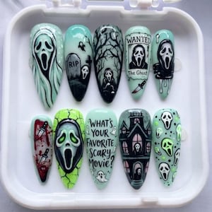 Op de afbeelding: Een set van tien amandelvormige press-on nagels met horror-thema in tinten mintgroen, zwart en wit. De kunstwerken bevatten het Ghostface-masker, een grafsteen met RIP, een spookhuis en de tekst What's Your Favorite Scary Movie?