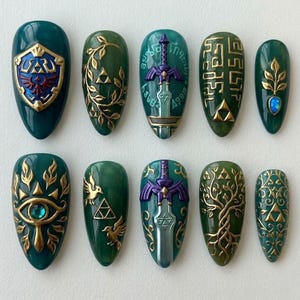 Peut inclure: Dix faux ongles vert &eacute;meraude avec des d&eacute;tails dor&eacute;s et bleus. Chaque ongle pr&eacute;sente un design diff&eacute;rent, dont un bouclier, une &eacute;p&eacute;e et divers symboles. Les ongles sont de forme ovale et ont une finition brillante.