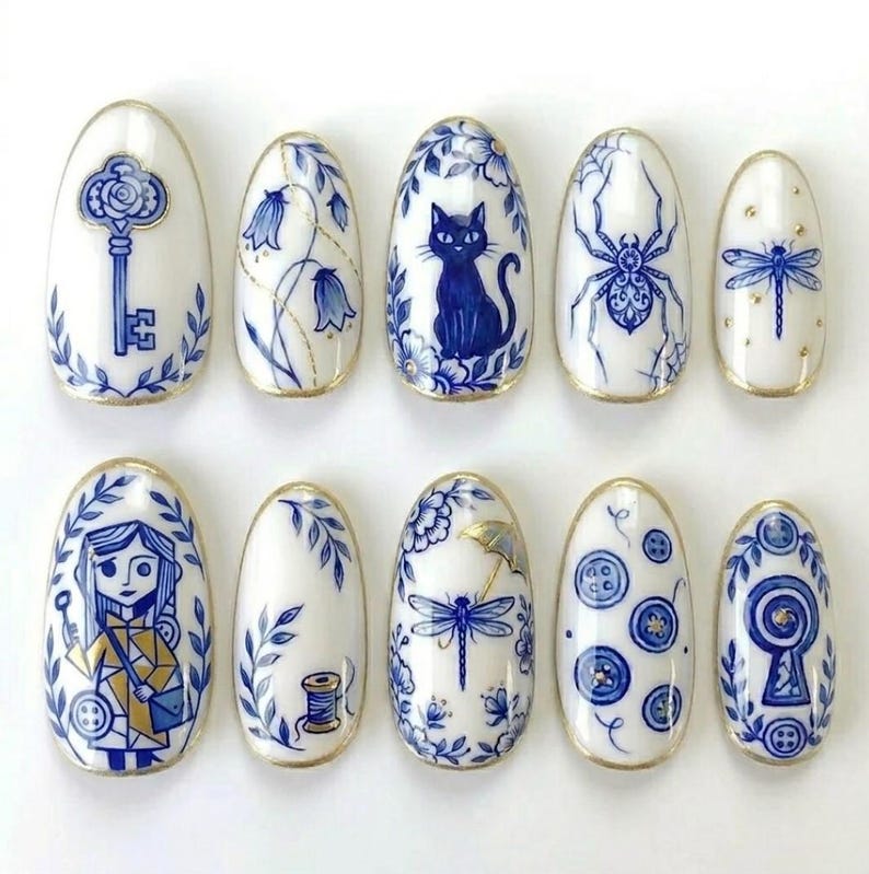 May include: A set of ten oval-shaped, white artificial nails with intricate blue and gold designs. The designs include a key, flowers, a cat, a spider, a dragonfly, a girl, a spool of thread, buttons, and a keyhole.