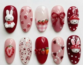 May include: A collection of ten artificial nails with various designs. The nails feature a range of colors, including red, pink, and white, and are decorated with strawberries, bows, hearts, and a bunny. Some nails have 3D embellishments.