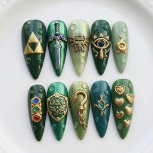 May include: A set of ten almond-shaped artificial nails in various shades of green. Each nail features a different design, including gold and silver embellishments of a sword, a shield, and other iconic symbols.