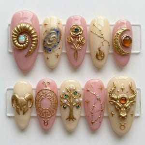 May include: A set of ten almond-shaped artificial nails with intricate designs. The nails feature a variety of gold-colored embellishments, including zodiac symbols, celestial patterns, and floral motifs, set against a backdrop of pale pink and cream colors.