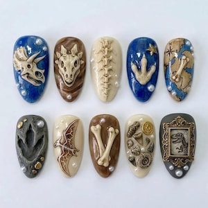 May include: A set of ten artificial nails with dinosaur-themed designs. Each nail features a different design, including dinosaur skulls, bones, footprints, and a map. The nails are painted in various colors, including blue, brown, and beige, and are adorned with pearls.