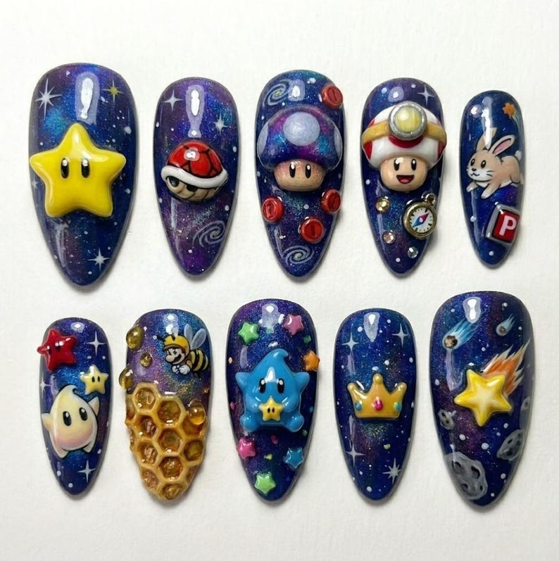 Pode incluir: Um conjunto de dez unhas artificiais com um design de tema gal&aacute;xia. Cada unha apresenta um personagem ou elemento 3D diferente, incluindo estrelas, um cogumelo, uma tartaruga, um coelho e uma abelha. As unhas s&atilde;o pintadas em tons de azul, roxo e branco.