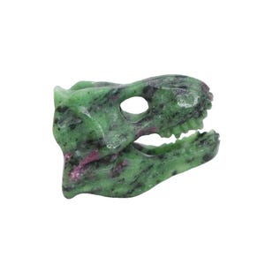 May include: A carved stone dinosaur skull in shades of green and pink. The skull features detailed teeth and eye sockets, showcasing the natural patterns of the stone. The sculpture is set against a plain white background.