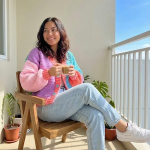May include: A multi-coloured crochet cardigan in pink, purple, blue, and orange. The open-front sweater has a round neckline and long sleeves. The person is wearing light blue jeans and white trainers. The person is sitting on a wooden chair.