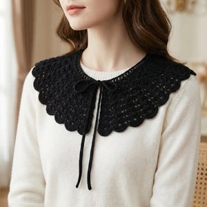 Könnte beinhalten: Ein schwarzer Häkelkragen mit gewelltem Rand und Bindeband. Der Kragen wird über einem weißen Pullover getragen. Das Lochmuster des Kragens verleiht dem Kleidungsstück eine zarte Note.