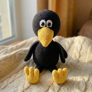 May include: A black crochet bird with a yellow beak and feet, and white-rimmed eyes. The handmade plush toy is sitting on a cream-coloured blanket. The bird has small black wings and is a cute, whimsical design.
