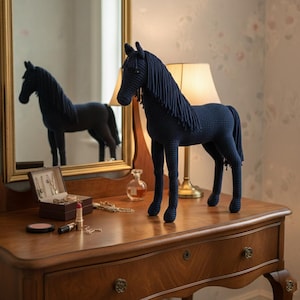 Puede incluir: Una figura de caballo de ganchillo azul marino está sobre una cómoda de madera. El caballo tiene una larga melena y cola. Un espejo refleja el caballo y la cómoda. Maquillaje y joyas están sobre la cómoda.