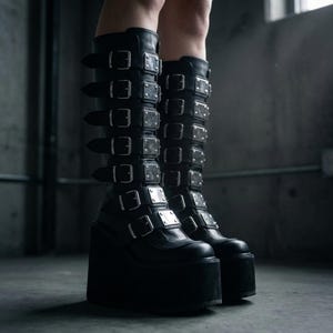 May include: Black platform boots with multiple buckles and silver-toned square accents. The boots have a chunky platform sole and extend to the knee. The boots are made of a smooth, dark material.
