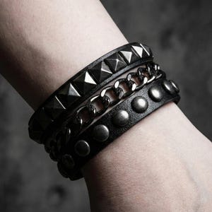 May include: Three black leather bracelets with metal accents. One bracelet features pyramid studs, another a chain, and the third has round studs. The bracelets are stacked on a wrist, showcasing a dark, edgy style.
