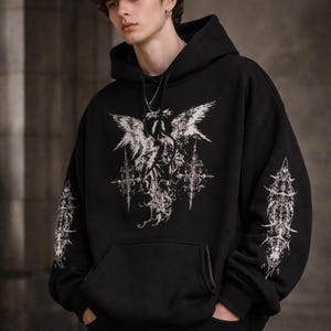 May include: Black hooded sweatshirt with a large white graphic on the front featuring an angel with outstretched wings and ornate crosses. The sleeves have matching white designs. The sweatshirt has a front pocket and a drawstring hood.