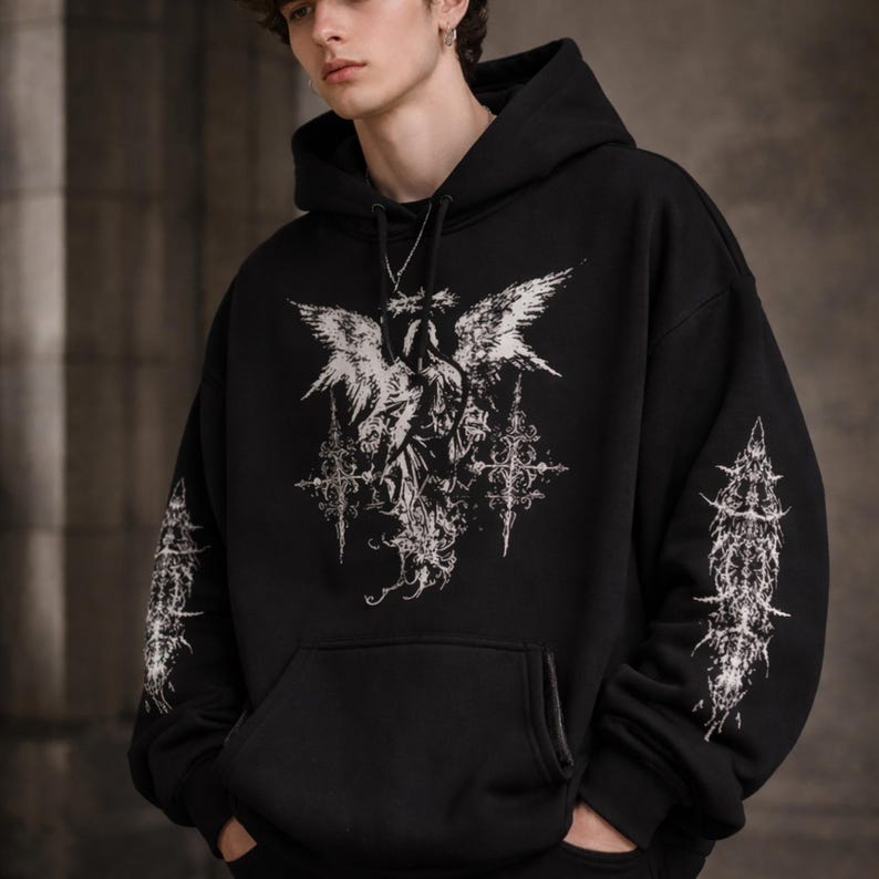 May include: Black hooded sweatshirt with a large white graphic on the front featuring an angel with outstretched wings and ornate crosses. The sleeves have matching white designs. The sweatshirt has a front pocket and a drawstring hood.