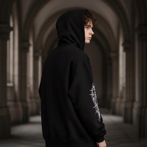 May include: Black hooded sweatshirt with a white graphic design on the left sleeve. The sweatshirt has a drawstring hood and a relaxed fit. The person in the image is wearing the sweatshirt in a dark, arched hallway.