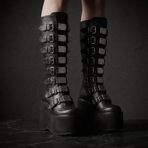 May include: A pair of black knee-high platform boots. Each boot features multiple silver-toned rectangular buckles and straps. The boots have a thick platform sole and a dark background.