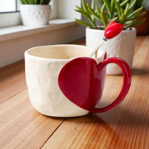 May include: Cream-colored ceramic mug with embossed heart pattern and a large red heart-shaped handle. A matching red heart-topped spoon rests inside the mug. The mug is on a wooden surface, with plants in the background.