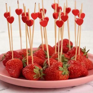 Puede incluir: Fresas frescas en un plato rosa, cada una ensartada con un palillo de madera rematado con un corazón rojo. Las fresas maduras y rojas tienen hojas verdes. Los palillos están espaciados uniformemente, creando una presentación festiva.