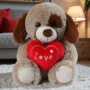 Può includere: Un peluche a forma di cane con pelo marrone e beige, che tiene un cuscino rosso a forma di cuore. Il cuscino ha la parola "Love" ricamata in bianco. Il cane ha occhi scuri e una macchia marrone sopra un occhio.