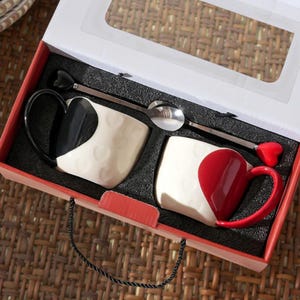 May include: A gift set featuring two ceramic mugs with heart-shaped designs, one black and white, the other red and white. Each mug has a matching spoon. The set is presented in a red box with a clear window, perfect for gifting.