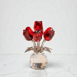 May include: A decorative crystal vase with a faceted design holds a bouquet of five red crystal roses. The roses have silver stems and are arranged in a silver leaf-shaped holder. The vase sits on a white marble surface.