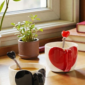 May include: Two ceramic mugs with heart designs. One mug has a large red heart, the other a black heart. Each mug has a matching heart-shaped spoon. A small potted plant and books are in the background.