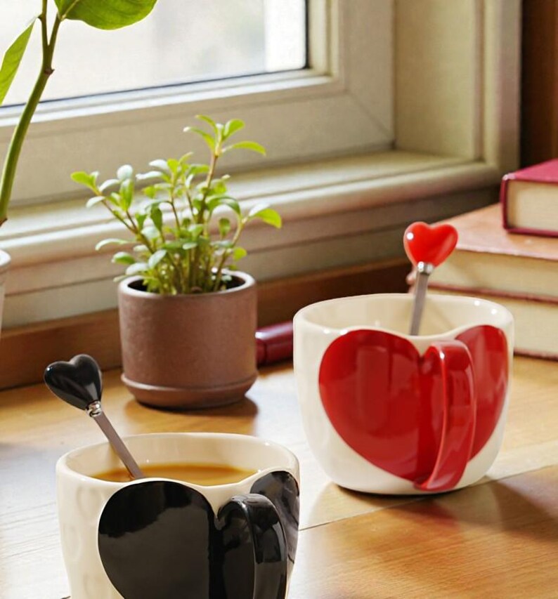 May include: Two ceramic mugs with heart designs. One mug has a large red heart, the other a black heart. Each mug has a matching heart-shaped spoon. A small potted plant and books are in the background.