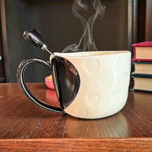 May include: A white ceramic mug with embossed heart shapes and a black heart-shaped handle. A matching black spoon rests inside the mug, which is filled with a hot beverage, emitting steam. The mug sits on a wooden surface.