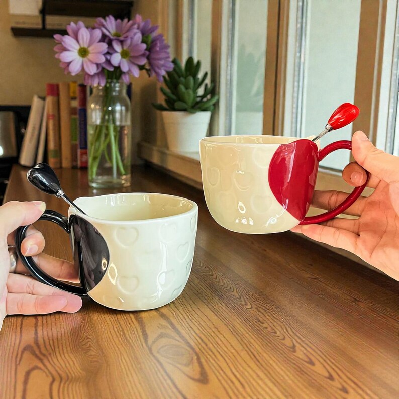 May include: Two ceramic mugs with heart-shaped and apple-shaped designs. One mug has a black handle and a black heart design, and the other has a red handle and a red apple design. Each mug has a matching spoon.
