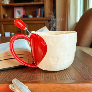 May include: A white ceramic mug with a red heart design and handle, holding a steaming beverage. A red spoon rests inside the mug. The mug is on a wooden table next to an open book.