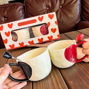 May include: A set of two ceramic mugs with heart-shaped designs and matching spoons. One mug has a black handle and a black heart design, while the other has a red handle and a red heart design. The mugs are presented in a box with heart patterns and the text "SWEET CUP".