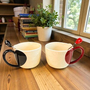 May include: Two cream-colored ceramic mugs with heart-shaped patterns. One mug has a black handle and a black heart-shaped design, the other has a red handle and a red heart-shaped design. Each mug has a spoon.