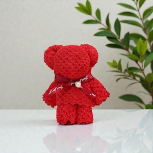 May include: A red teddy bear made from a towel, with a dark red ribbon tied around its neck. The ribbon has the text "Just for you" printed on it. A white pearl is attached to the ribbon. The bear is standing upright.