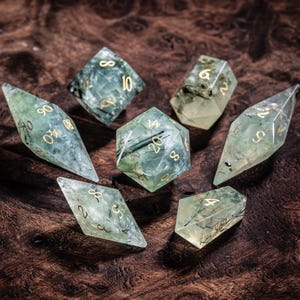 May include: A set of seven polyhedral dice made from translucent pale green resin with dark needle-like inclusions and engraved gold-colored numbers. The collection includes various geometric shapes such as a twenty-sided die, a twelve-sided die, and several diamond-shaped dice, all featuring sharp edges and clear, polished surfaces.