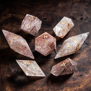 May include: A set of six polyhedral dice crafted from clear material with embedded reddish-brown needle-like inclusions. Each die features engraved numbers filled with gold-coloured ink, including a twenty-sided die, a percentile die, and various others.