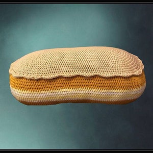 May include: A crocheted pillow in the shape of a long loaf of bread. The pillow is made with a beige yarn and has a yellow and white striped pattern.