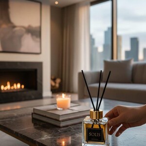 Könnte beinhalten: Ein klarer Glasdiffusor mit goldenem Deckel und schwarzen Stäbchen, beschriftet mit "SOLIS", steht auf einem Marmor-Couchtisch. Eine brennende Kerze und Bücher befinden sich ebenfalls auf dem Tisch. Der Hintergrund umfasst einen Kamin, ein Sofa und eine Stadtansicht.