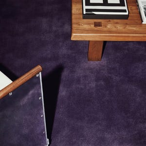 May include: A wooden coffee table with a black and white book on top, positioned on a deep purple carpet. A chair with a wooden armrest and a purple upholstered side panel is visible in the foreground, featuring silver metal rivets along the edge.