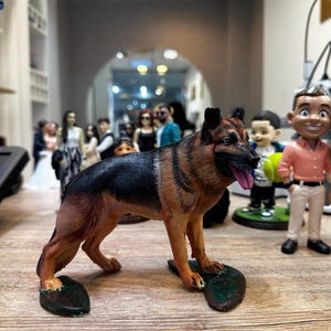 May include: A detailed figurine of a German Shepherd dog, featuring black and tan fur, a pink tongue, and a realistic pose. The dog stands on a green base. Other figurines are visible in the background.