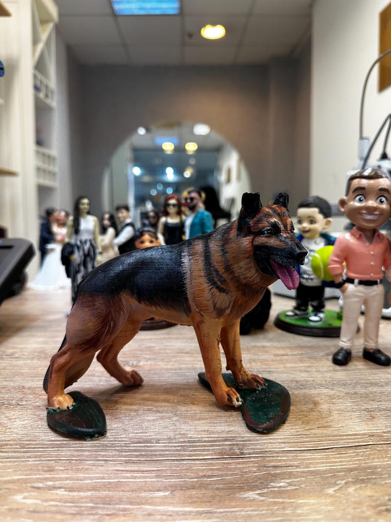 May include: A detailed figurine of a German Shepherd dog, featuring black and tan fur, a pink tongue, and a realistic pose. The dog stands on a green base. Other figurines are visible in the background.