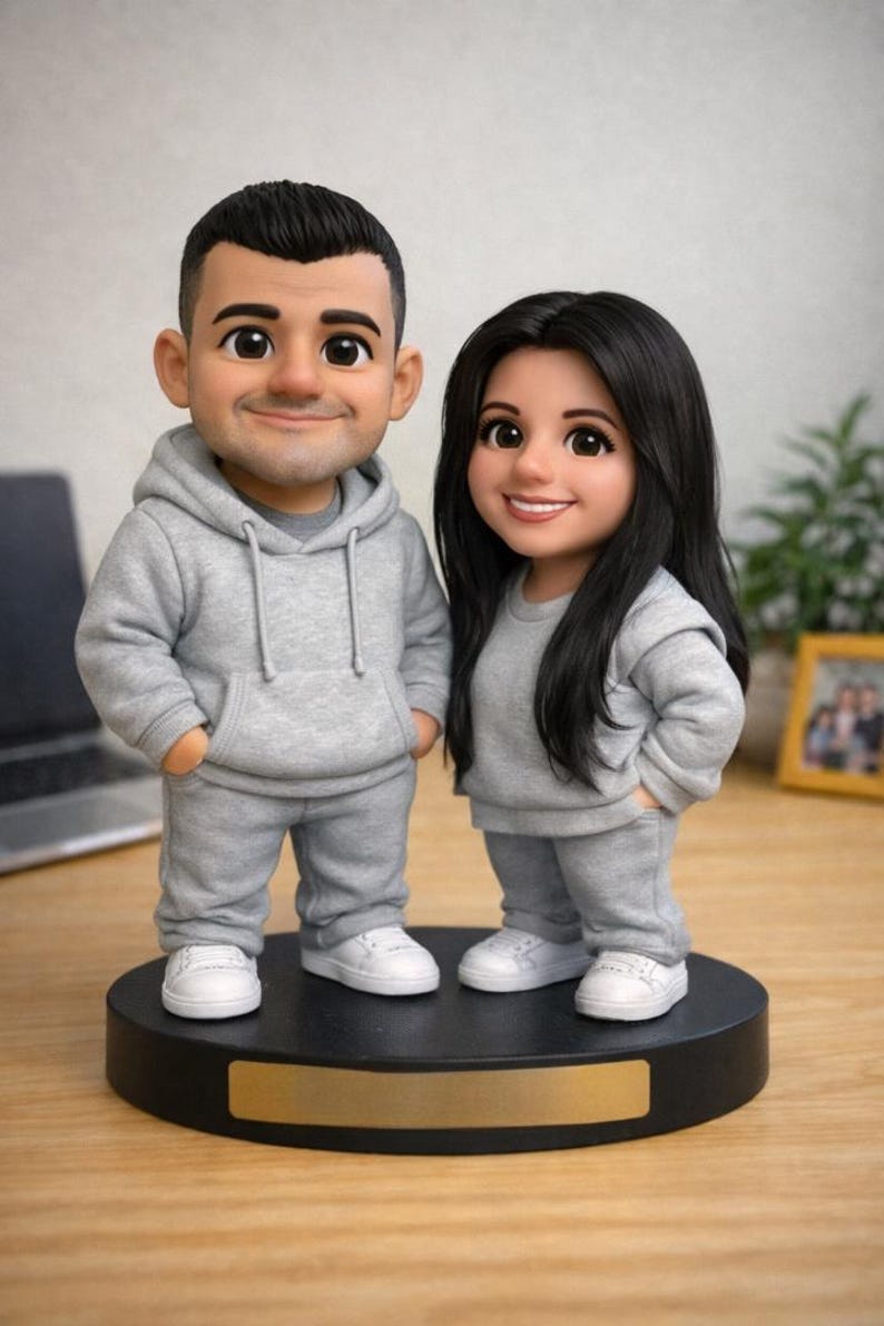May include: A figurine of a couple, both wearing gray sweat suits and white sneakers, standing on a black base. The male figure has short black hair, and the female figure has long black hair. The base has a gold-colored nameplate.