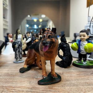 May include: A detailed figurine of a German Shepherd dog, featuring brown and black fur, a pink tongue, and a green base. Other figurines are visible in the background, including wedding cake toppers and a child holding tennis balls.