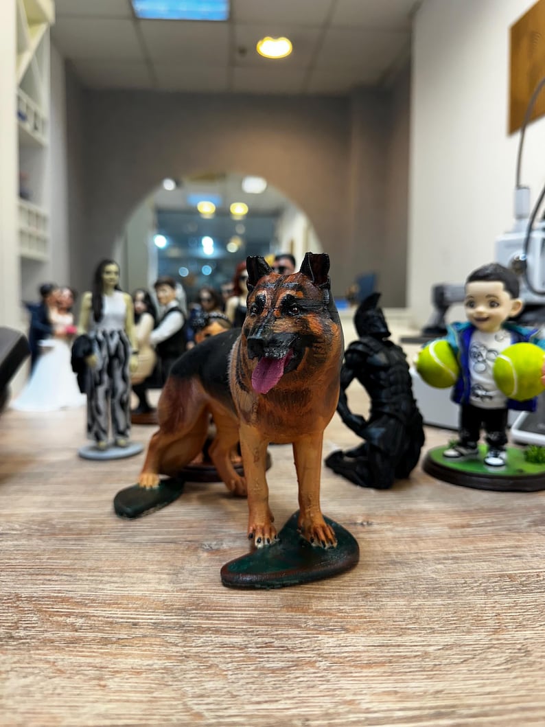 May include: A detailed figurine of a German Shepherd dog, featuring brown and black fur, a pink tongue, and a green base. Other figurines are visible in the background, including wedding cake toppers and a child holding tennis balls.