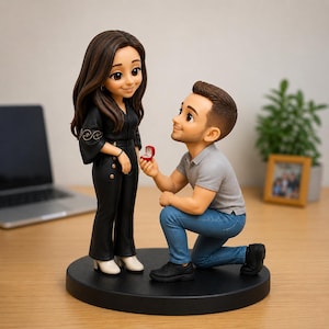 Peut inclure: Une figurine personnalisée représentant une personne aux longs cheveux bruns vêtue d'une combinaison noire debout, tandis qu'une autre aux cheveux bruns courts en polo gris et jean bleu est à genoux, tenant une petite boîte à bague rouge.