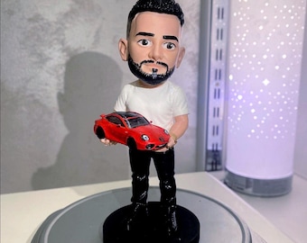 May include: A bobblehead figurine of a person with a beard, wearing a white t-shirt and black pants, holding a small red sports car. The figure stands on a black circular base with the yellow text 10 Jahre Jubiläum printed on the front edge.